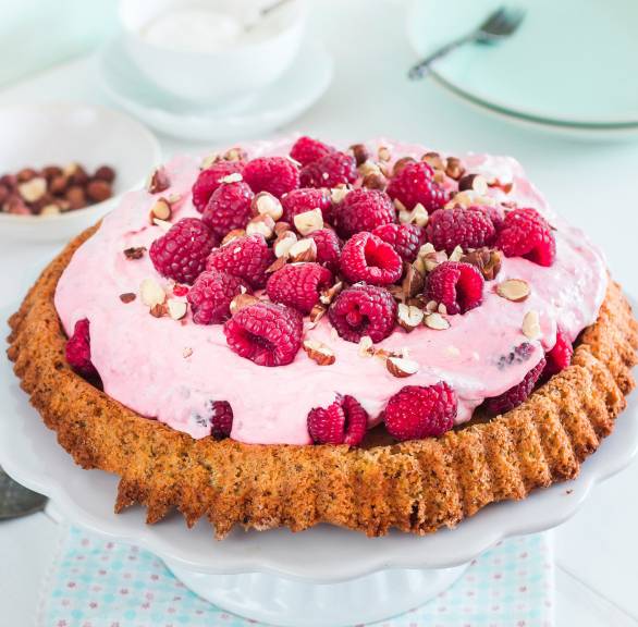 Selbstgebackener Haselnusskuchen getoppt von mit Himbeer-Mascarponecreme, Himbeeren und Haselnusskernen auf einer Kuchenplatte