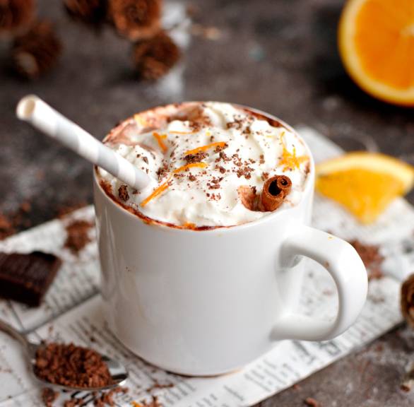 Heiße Schokolade mit Sahne in einer Tasse auf einem Tisch, daneben Zimtstangen und Orangen