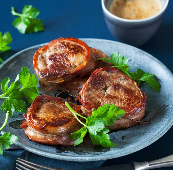 Drei Kalbsmedaillons umhüllt mit Parmaschinken auf einem Teller, daneben Sahnesoße in einer Schale