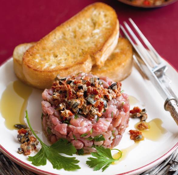 Kalbstartar mit Tomaten-Oliven-Topping auf einem Vorspeisenteller angerichtet, daneben eine Scheibe geröstetes Ciabatta