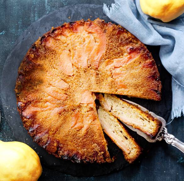 Runder Quittenkuchen angeschnitten auf einer Kuchenplatte, daneben zwei Quitten auf einem Tisch