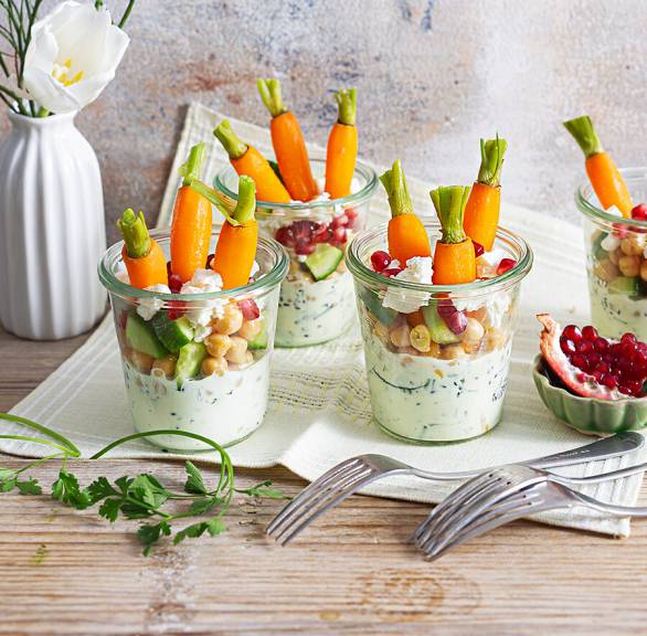 Schichtsalat mit Karotten, Kichererbsen, Feta, Joghurtdressing und Granatapfelkernen in vier Gläsern auf einem festlich dekorierten Tisch 