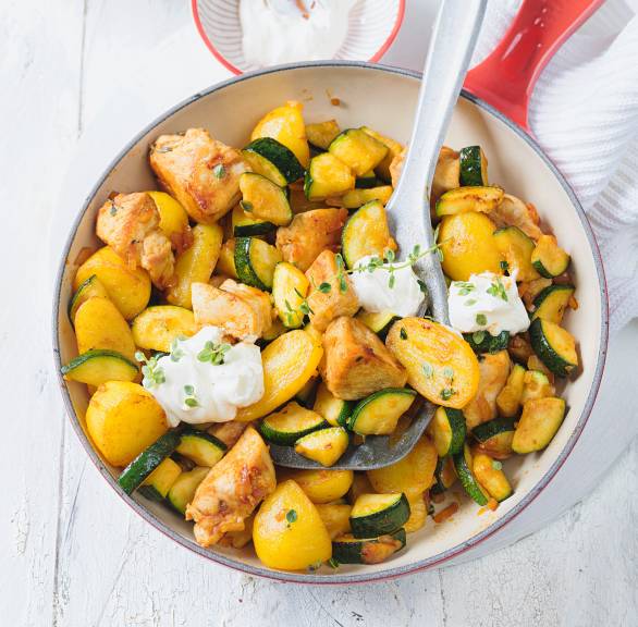 Pfanne mit Hähnchen, Kartoffeln und Zucchini getoppt von Schmand-Klecksen und frischen Kräutern auf einem Tisch