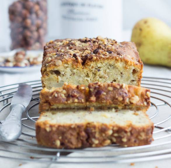 Angeschnittener Kartoffelkuchen aus der Kastenform auf einem Kuchengitter, daneben Haselnüsse und Birnen auf einem Tisch