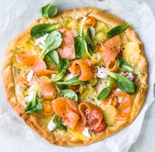 Runde Kartoffelpizza belegt mit Räucherlachs, Tomaten und Feldsalat auf einem Stück Backpapier