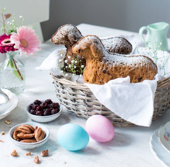 Zwei Osterlämmer in einem Korb, daneben Nüsse und Cranberries sowie Ostereier auf einem dekorierten Tisch