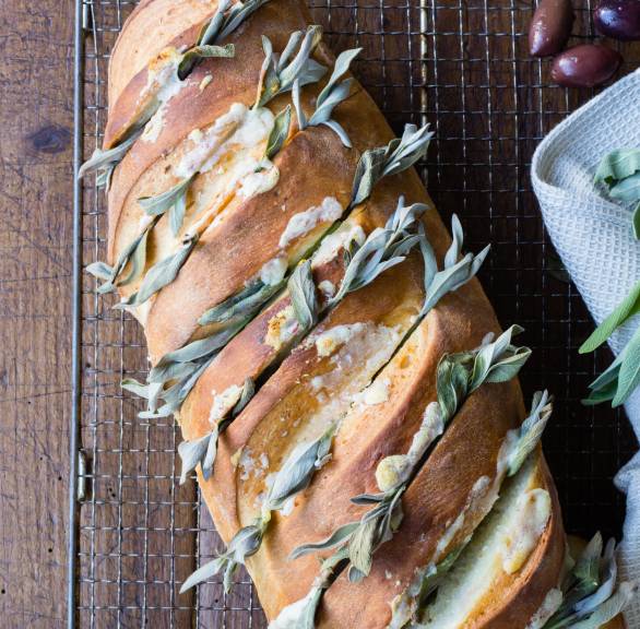Selbst gebackenes Knoblauchbrot eingeschnitten und gefüllt mit Knoblauch, Parmesan und dekoriert mit Salbeiblättern auf einem Holztisch