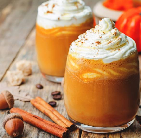 Kürbis-Caffè-Latte in zwei Gläsern getoppt mit Sahnehaube, daneben Zimtstangen