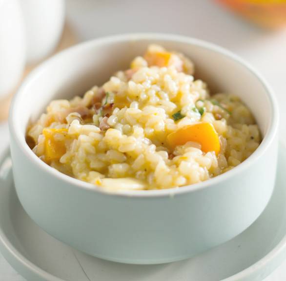Kürbisrisotto in einer Schale auf einem Tisch, dahinter ein Stück Hokkaidokürbis