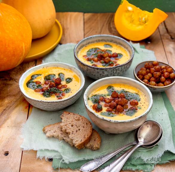 Selbstgekochte Muskatkürbis-Suppe in drei Supperschalen getoppt mit Kichererbsen und Kräuteröl auf einem Holztisch