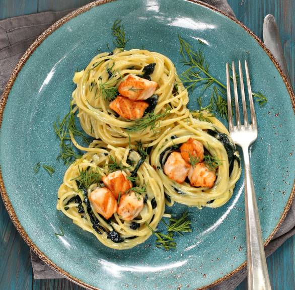 Drei Spaghettinester auf einem blauen Teller getoppt von Lachswürfeln und Spinat