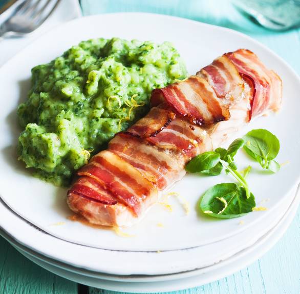 Ein Stück Lachsfilet im Speckmantel auf einem Teller, daneben grüner Kartoffelstampf und frische Kresse