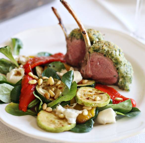Lammkarree mit Parmesankruste angerichtet auf Antipastisalat getoppt von Pinienkernen auf einem Teller