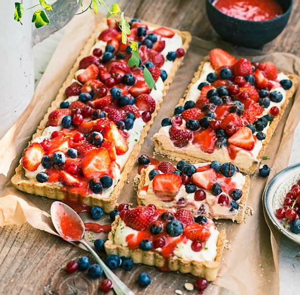 Zwei gebackene Beeren-Tartes mit Mascarpone-Joghurt-Creme getoppt von frischen Beeren angerichtet auf einem Holztisch