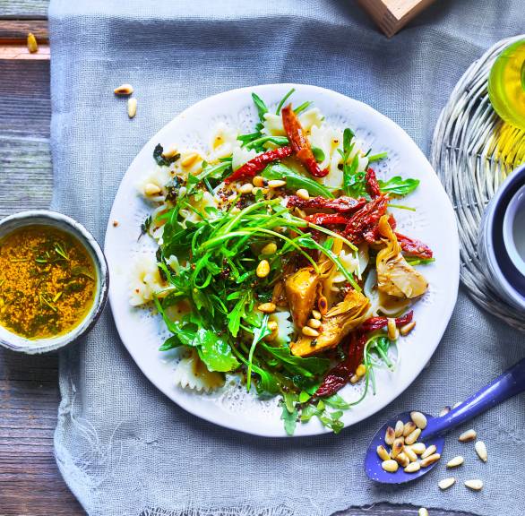 Mediterraner Nudelsalat auf einem Teller bestreut mit Pinienkernen, daneben eine Schale mit Vinaigrette