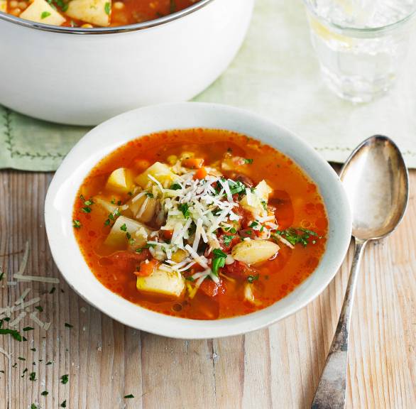 Minestrone in einer Suppenschüssel getoppt von Patmesanspänen, daneben ein voller Suppentop mit einer Kelle darin