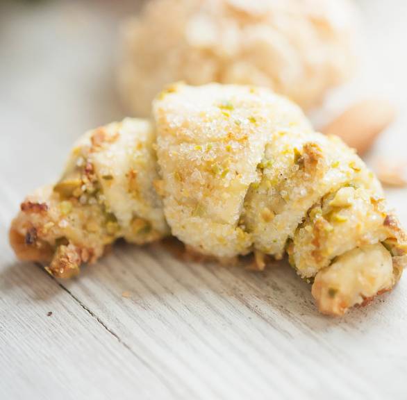 Ein Vanille-Hörnchen aus Frischkäseteig mit Pistazienfüllung und -topping auf einem hellen Holztisch