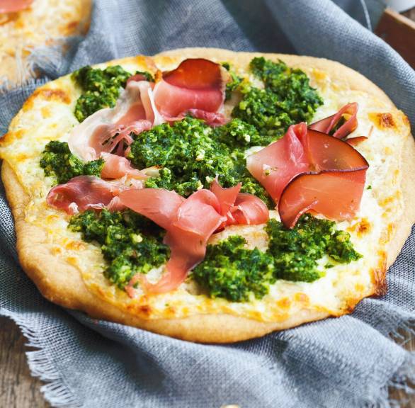 Zwei Minipizzen auf einem Holztisch mit Grünkohlpesto und Speck, daneben Walnusskerne