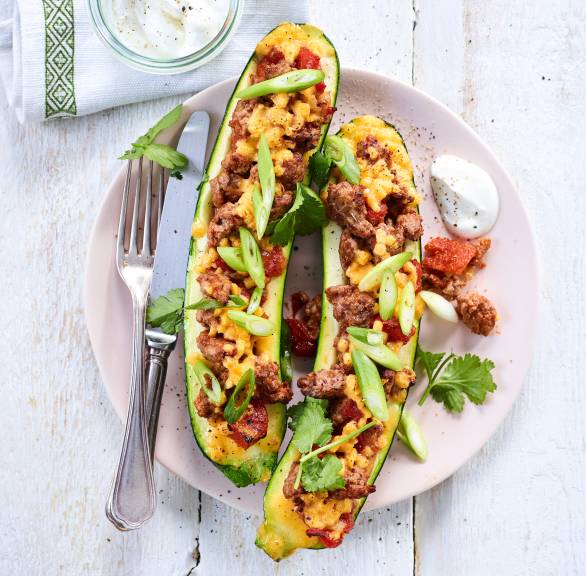 Zwei halbe, gebackene Zucchini gefüllt mit Hackfleisch à la Bolognese und mit Mozzarella überbacken angerichtet auf einem Teller mit Joghurtdip
