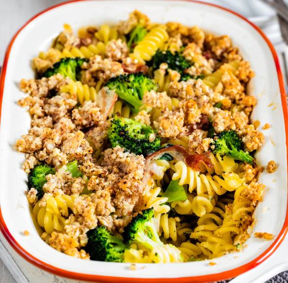 Nudelgratin mit Brokkoli, Pancetta und Mandelhaube in einer Auflaufform auf einem weißen Holztisch