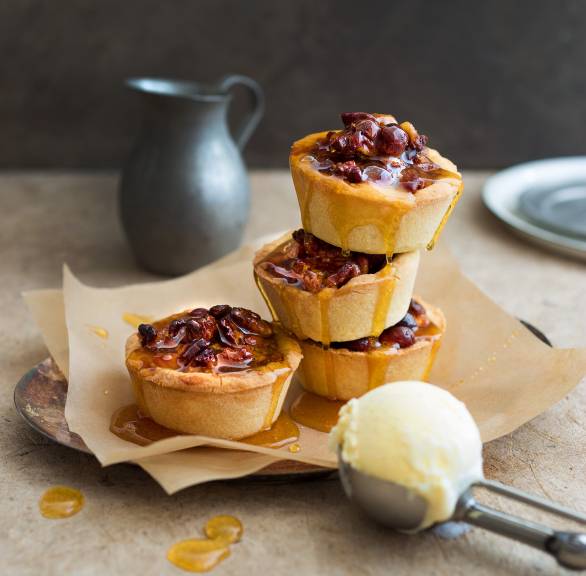 Vier karamellisierte Honig-Nuss-Törtchen mit Orangenguss angerichtet auf einem Dessertteller, daneben ein Eisportionierer mit einer Kugel Vanilleeis