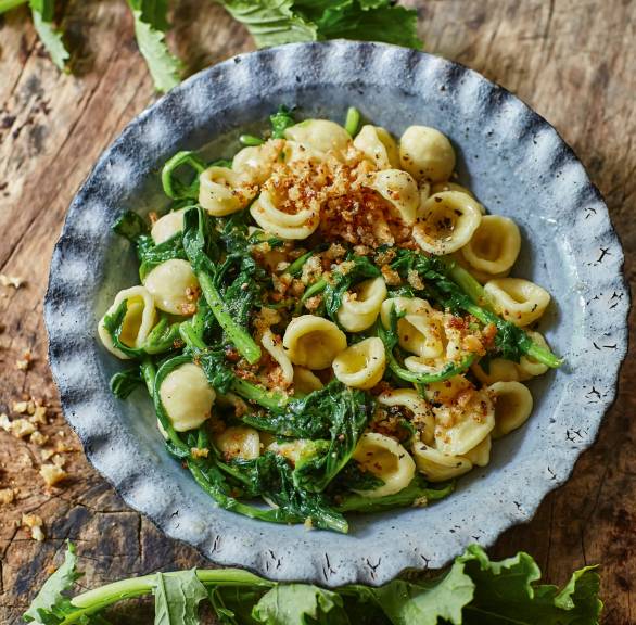 Orecchiette mit Cime di Rapa angerichtet auf einem Teller, daneben Stängelkohl auf dem Tisch