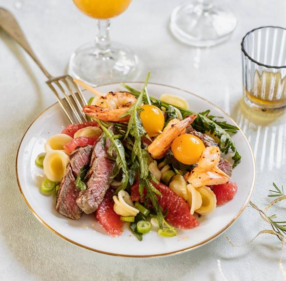 Nudelsalat mit Steakstreifen, Garnelenspießen und Grapefruitstücken auf einem Teller, daneben Dressing in einem Glas