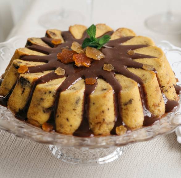 Italienischer Brotpudding aus Panettone steht auf einer Tortenplatte und ist mit Schokosoße übergossen
