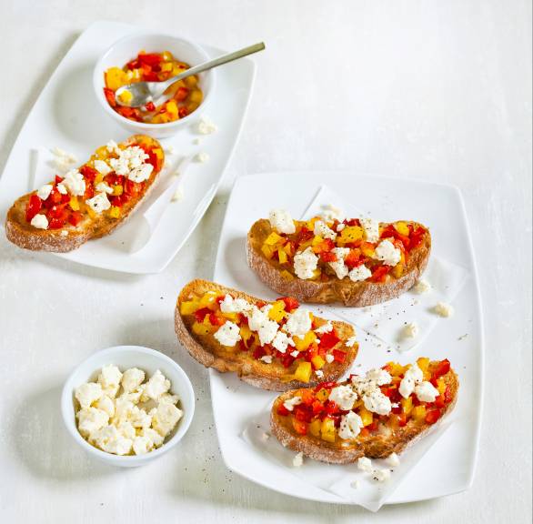 Bruschetta auf einem Teller getoppt mit Paprikawürfeln und Schafskäse, daneben Schalen mit Paprika und Schafskäse