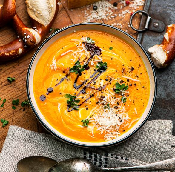 Kürbissuppe in einer Schüssel getoppt von Parmesan und Balsamico, daneben eine Laugenbrezel auf einem Tisch