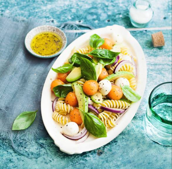 Pastasalat mit Melonenbällchen und Mozzarellakugeln auf einem ovalen Teller, darauf Avocadostücke