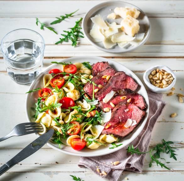 Teller mit Pastasalat auf der einen Seite und Rindfleischscheiben auf der anderen, daneben Parmesan und Pinienkerne auf einem Tisch