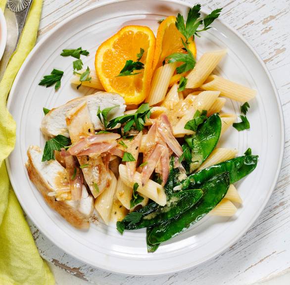 Hähnchenbrustfiletstreifen angerichtet mit Ricotta-Orangensauce, Penne, Zuckerschoten und Orangenscheiben auf einem Teller