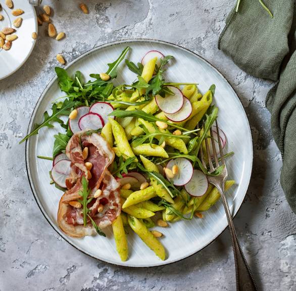 Penne-Salat auf einem Teller mit Pancettastreifen und Rucola, daneben grünes Pesto in einem Glas