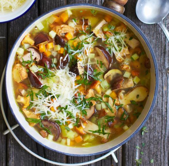 Klare Pilzsuppe mit Gemüse in einem Stieltopf auf einem Tisch, daneben gehobelter Käse und Baguette