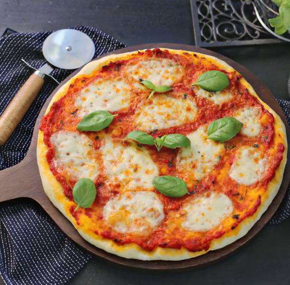 Runde, ganze Pizza Margherita auf einem Pizzabrett, daneben frische Basilikum in einem Topf