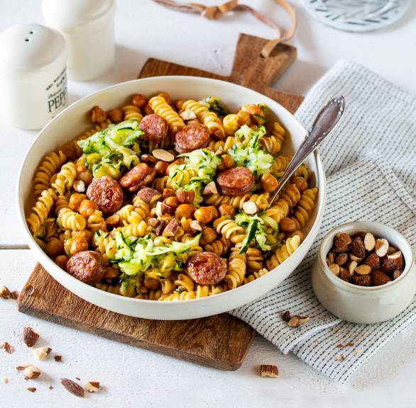 Riccioli Nudelsalat mit Kichererbsen, gebratener Salsiccia und Zucchiniraspeln angerichtet in einer Pastasalatschüssel, daneben Rauchmandeln