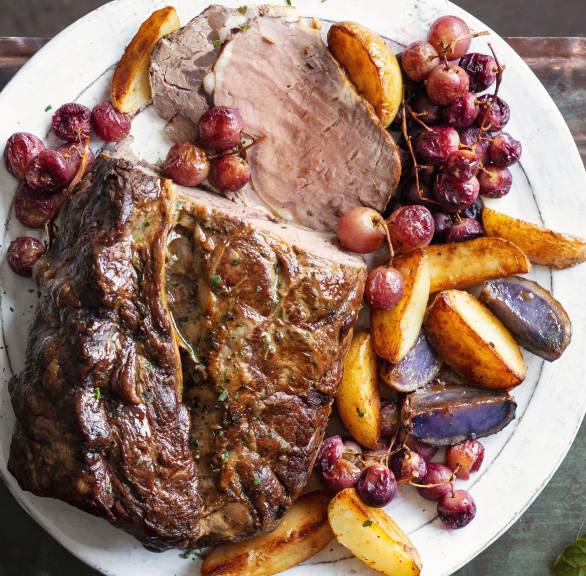 Gebackenes Roastbeef am Stück angeschnitten auf einem Teller umgeben von gebackenen Kartoffeln und Trauben