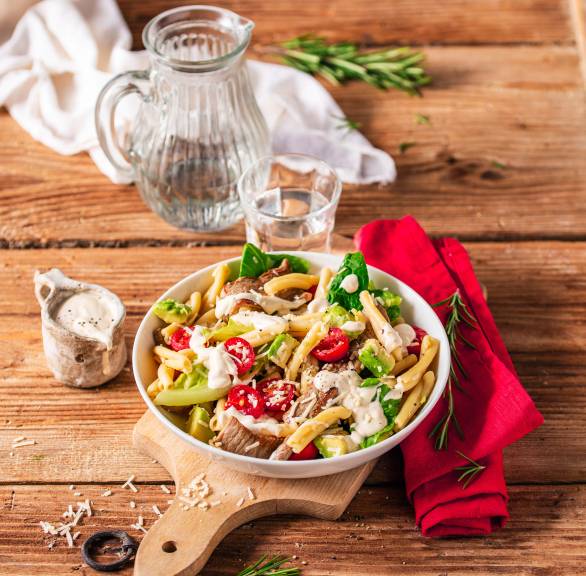 Pastasalat mit Romana auf einem Teller, getoppt von Kalbsschnitzelstreifen, Avocadostücken und einem cremigen Dressing
