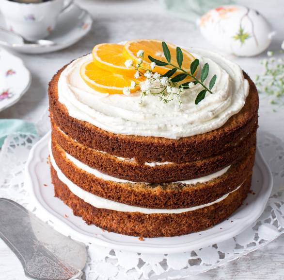 Rüblitorte mit Orangen-Frischkäse-Füllung getoppt von Orangenscheiben auf einer Tortenplatte auf einem österlich dekorierten Tisch