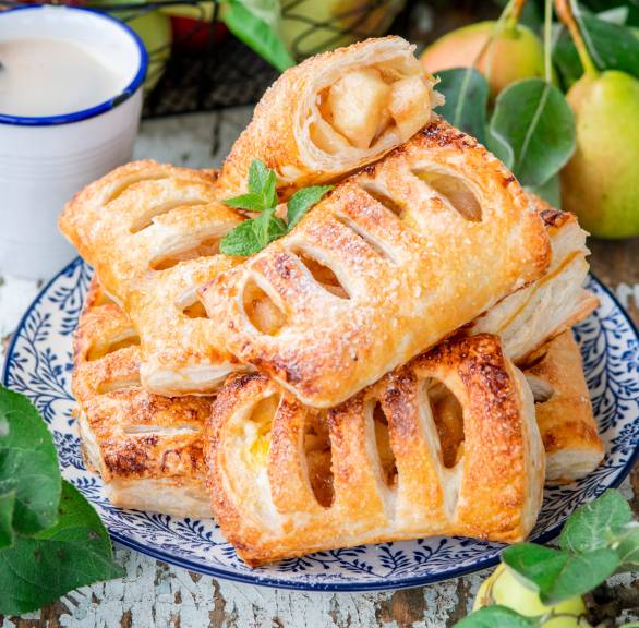 Bratapfeltaschen aus Blätterteig gestapelt auf einem Teller, daneben frische Äpfel auf einem Tisch
