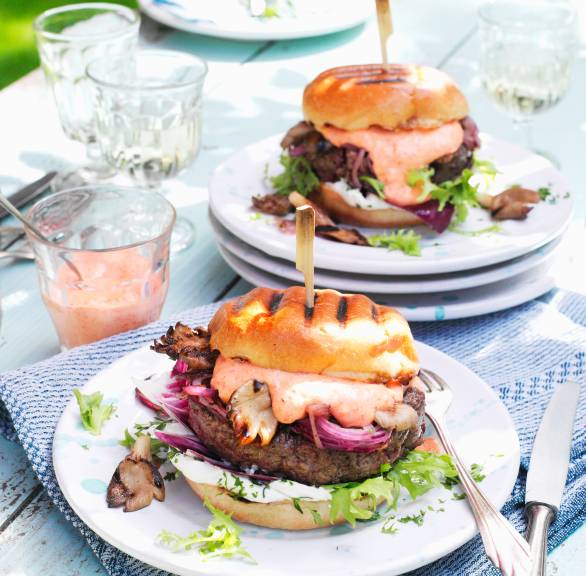 Gegrillter Burger mit Ziegenkäse und Pilzen gefüllt, zusammengehalten von einem Holzstab auf einem Teller, daneben ein Glas Mayonnaise