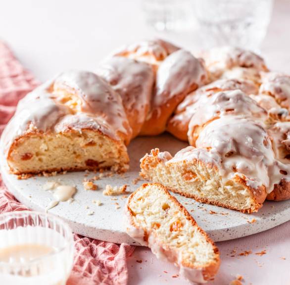 Osterzopf mit Guss angeschnitten auf einer Tortenplatte, daneben Sahne und getrocknete Aprikosen
