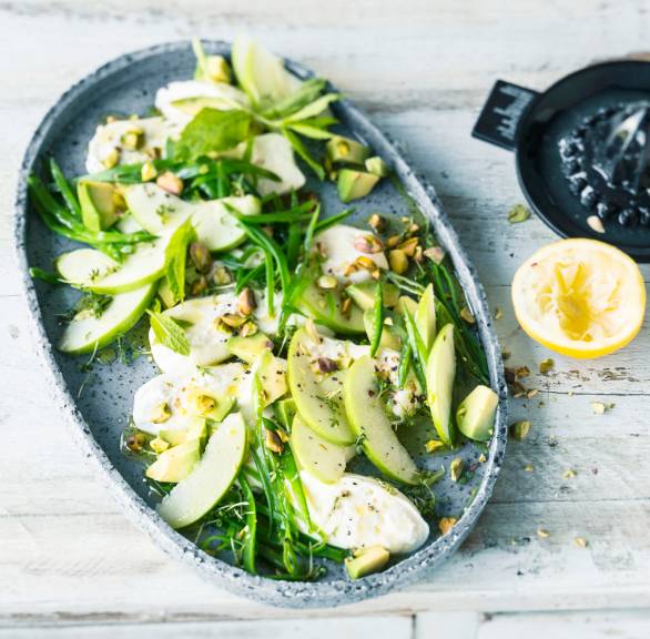 Burrata-Salat mit grünen Apfelspalten auf einem schwarzen Teller getoppt von Pistazien, daneben eine halbe Zitrone auf einem Tisch