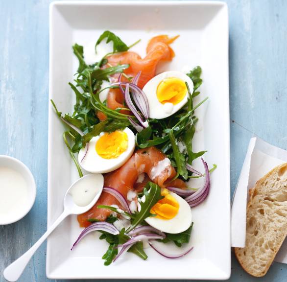 Rucola-Salat getoppt von Räucherlachsscheiben, halben gekochten Eiern und Joghurt-Dressing angerichtet auf einer Salatplatte