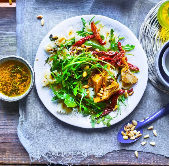 Farfalle-Salat angerichtet auf einem Teller mit Artischocken, Rucola, getrockneten Tomaten und Pinienkernen