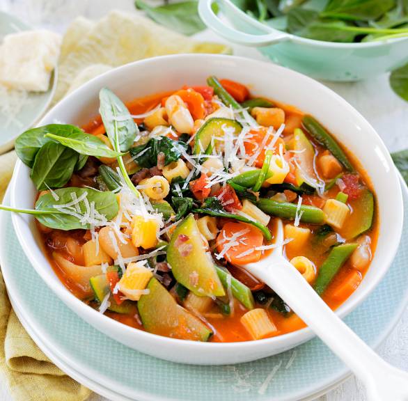 Sommer-Minestrone in einer Suppenschüssel, daneben Babyspinatblätter und Parmesan in Schalen