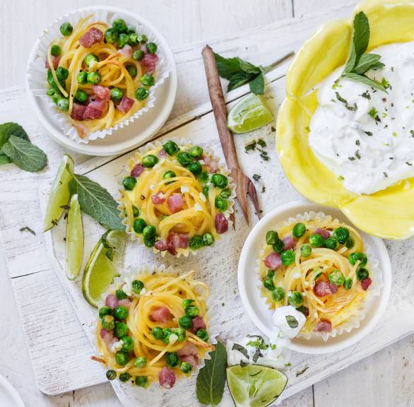 Mehrere Spaghetti-Muffins mit Schinken in papierförmchen auf einem Tisch, daneben Minz-Limetten-Dip