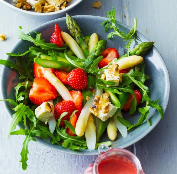 Spargelsalat mit Erdbeeren in einer Schüssel, daneben Joghurtdressing, Walnüsse und Feta auf einem Tisch