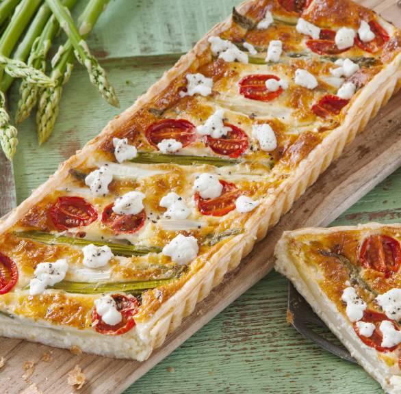 Reechteckige Spargeltarte mit Ziegenfrischkäse und Kirschtomaten angeschnitten auf einem Holzbrett